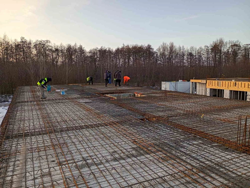 Robotnicy przygotowują zbrojenie płyty fundamentowej dużego budynku na tle drzew