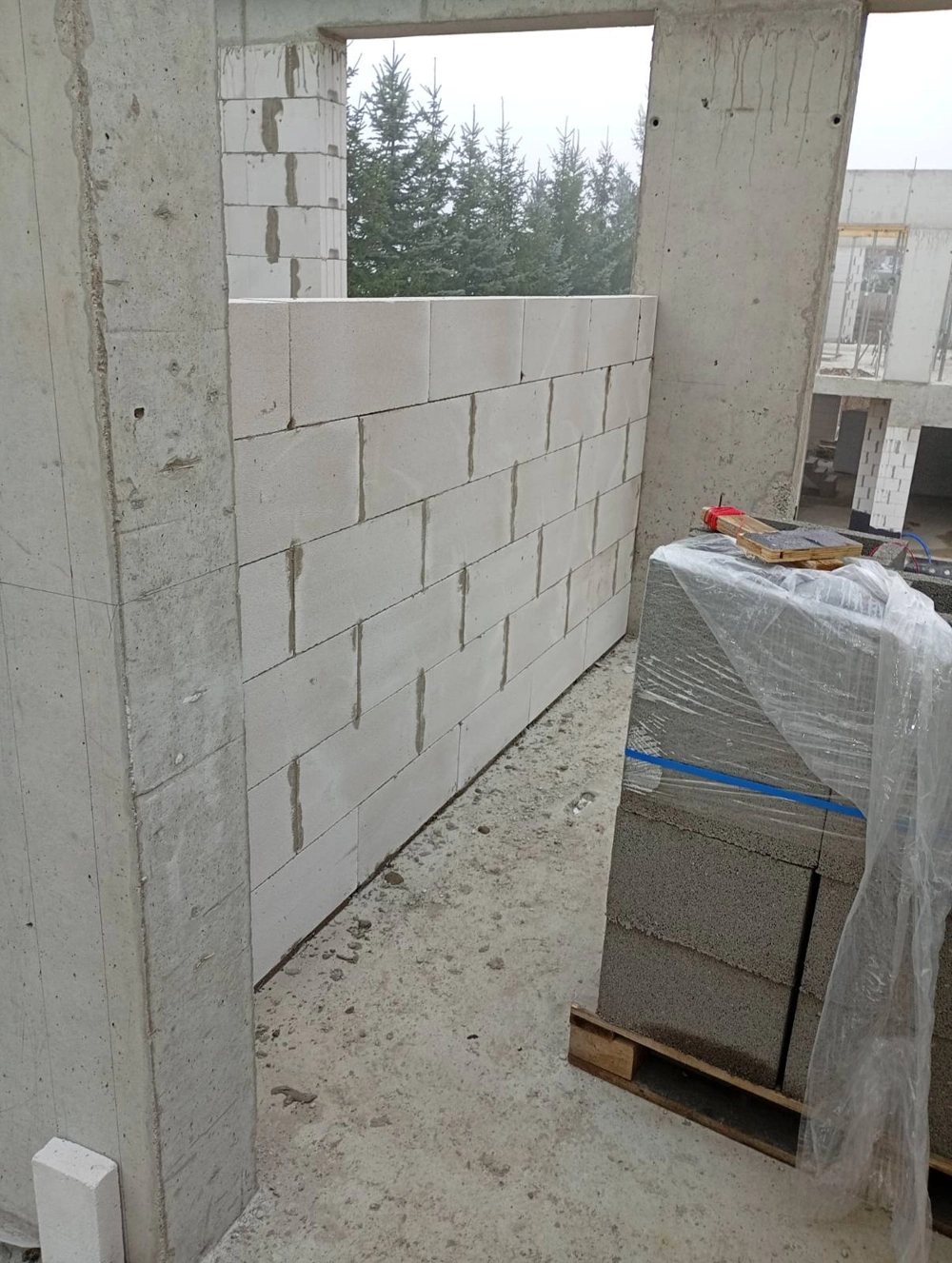 Budowa ściany działowej z białych bloczków na stropie betonowym, paleta bloczków i słupy żelbetowe w tle