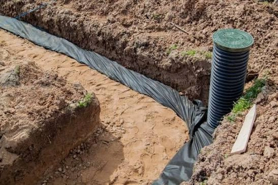 Instalacja drenażu opaskowego z czarną rurą drenarską i geowłókniną w wykopie