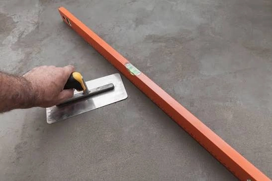 Ręka robotnika wygładza świeżą betonową wylewkę pacą obok poziomicy na podłodze Wylewki